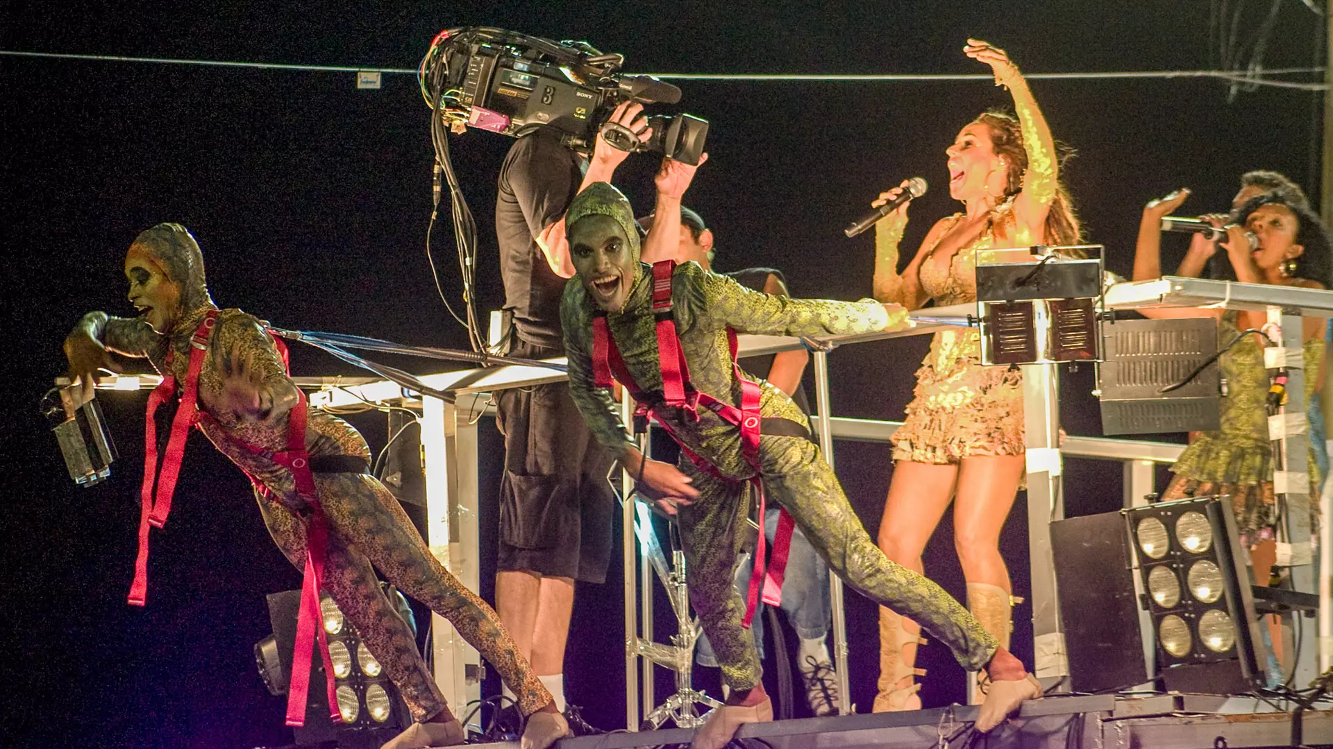 Cantores, dançarinos e câmera em cima de um trio elétrico durante o Carnaval de Salvador à noite.